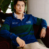 A man wearing a blue and green striped quarter-zip sweatshirt with white embroidered text, sitting in a dark red armchair in a cafe-like setting.