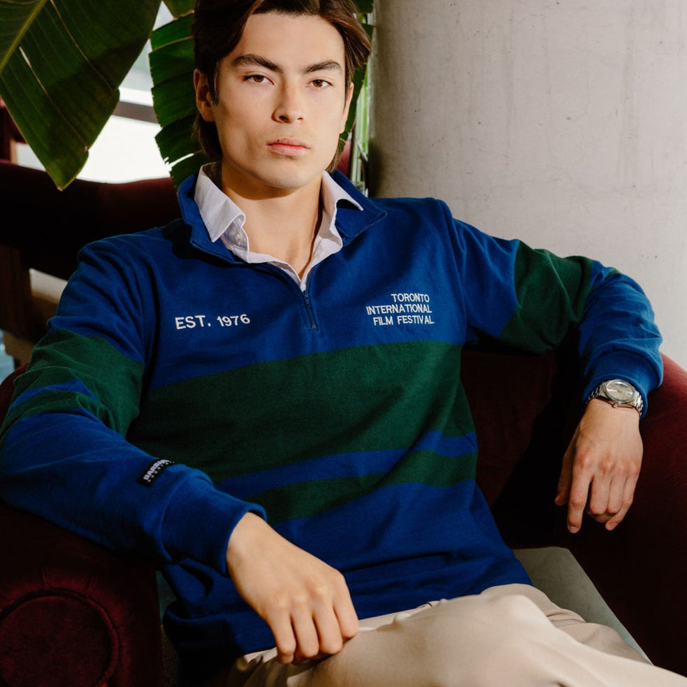 A man wearing a blue and green striped quarter-zip sweatshirt with white embroidered text, sitting in a dark red armchair in a cafe-like setting.