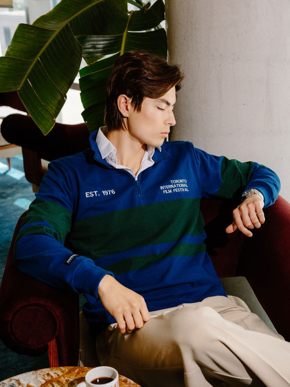 A man wearing a blue and green striped quarter-zip pullover sweater with 'EST. 1975' and 'TORONTO INTERNATIONAL FILM FESTIVAL' printed on the chest, sitting in a red armchair.