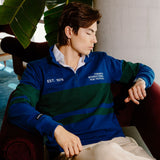 A man wearing a blue and green striped quarter-zip pullover sweater with 'EST. 1975' and 'TORONTO INTERNATIONAL FILM FESTIVAL' printed on the chest, sitting in a red armchair.