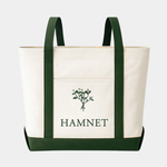 A cream-colored canvas tote bag featuring dark green vertical straps and a matching base, with a central pocket displaying a green botanical illustration above the word "HAMNET" in bold, green serif letters.