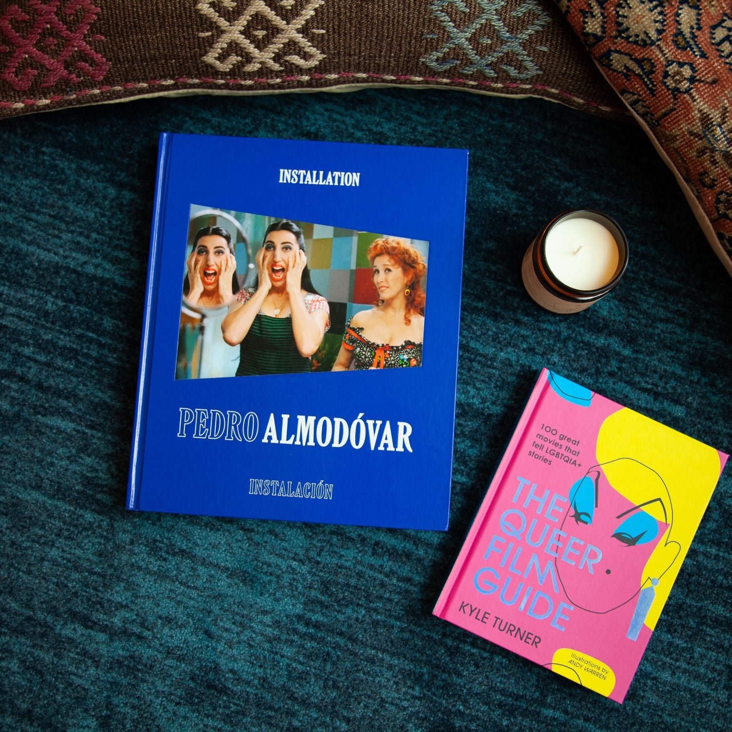 Two books, one a large blue volume titled "Pedro Almodóvar: Installation" and the other a pink and yellow book titled "The Queer Film Guide," sit on a textured teal rug alongside a small lit candle and patterned pillows.