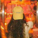 Person wearing a yellow 'POP CORN' cap in front of a popcorn machine.