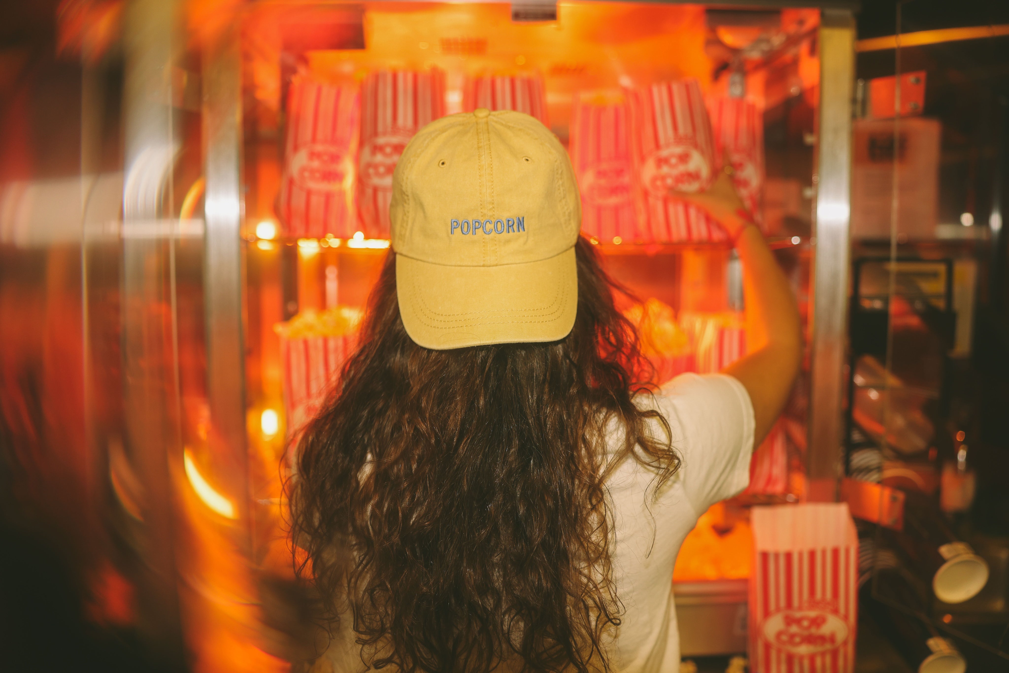 Person wearing a yellow 'POP CORN' cap in front of a popcorn machine.
