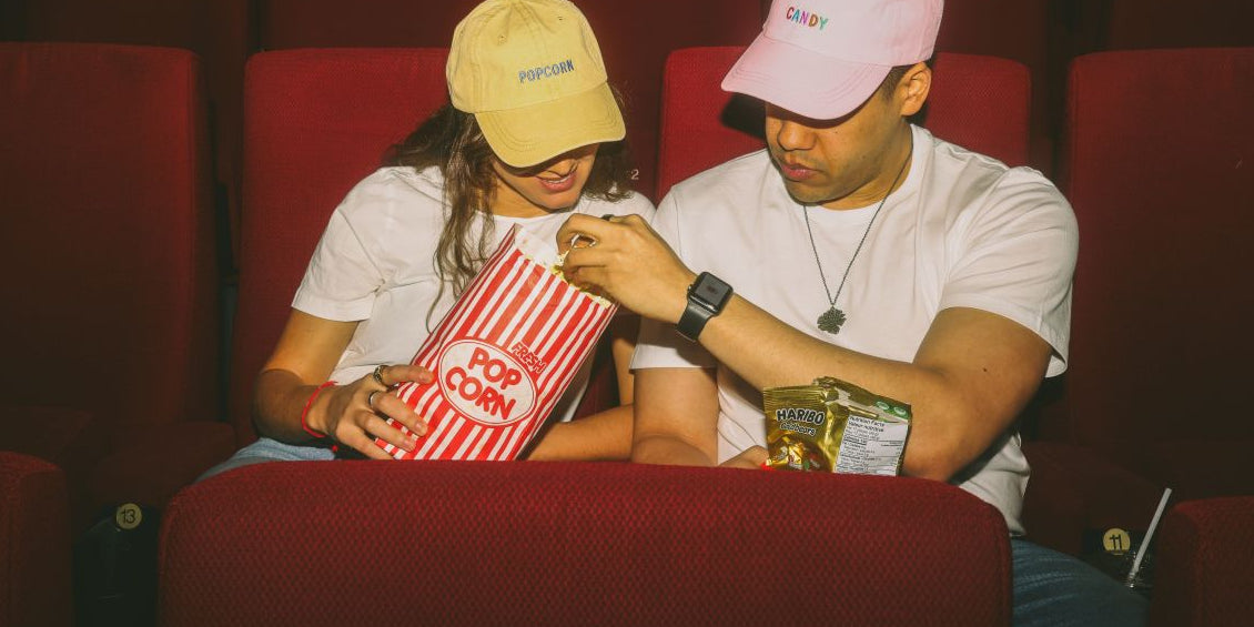 A man and a woman wearing baseball caps sit in a movie theater together, sharing a large bag of popcorn and a package of gummy bears.