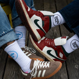 Two people sit on a wooden surface with their feet overlapping, showcasing two pairs of Air Jordan sneakers in a red, white, and green colorway, one pair of brown Adidas sneakers, and white crew socks that read "Inside Out."
