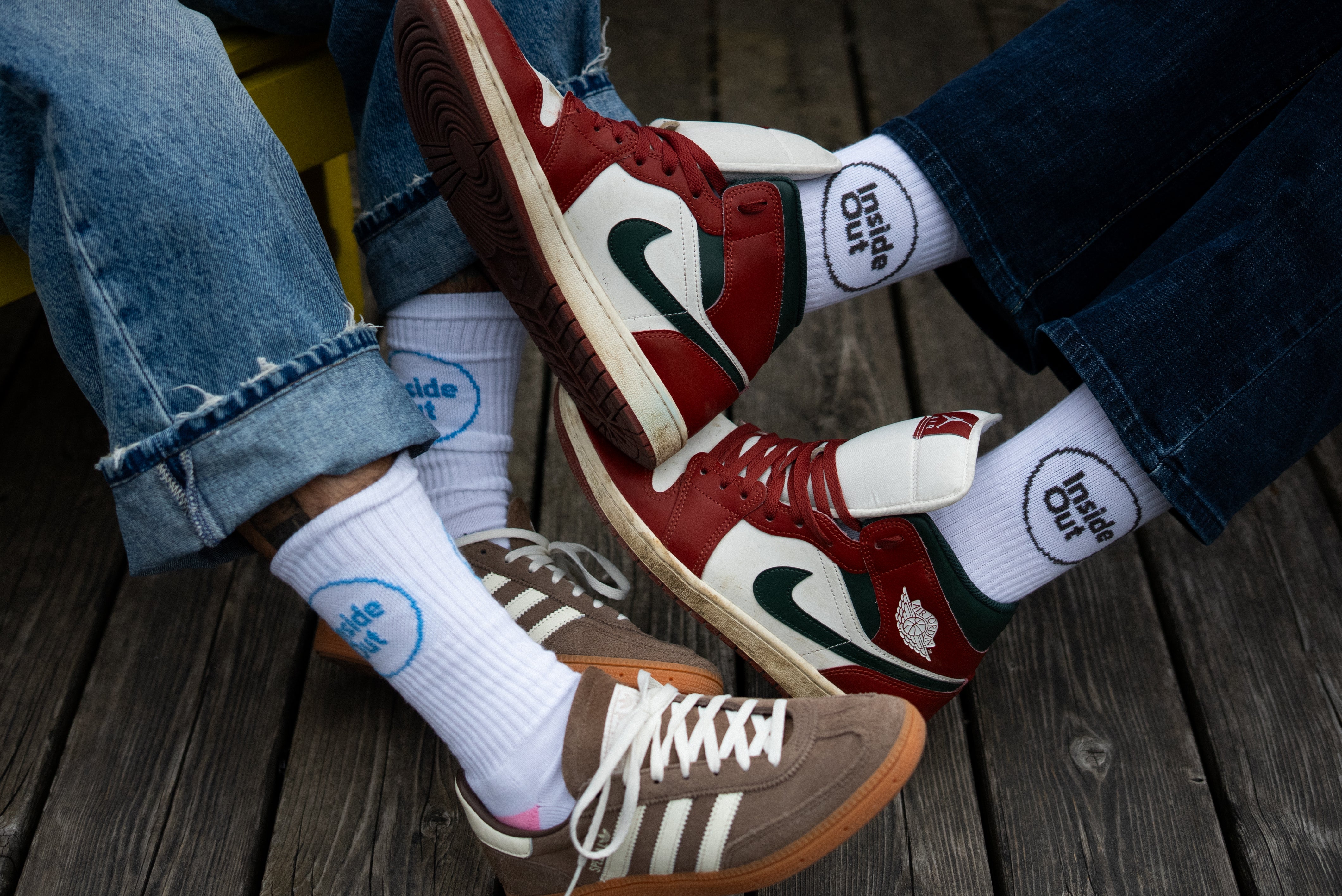 Two people sit on a wooden surface with their feet overlapping, showcasing two pairs of Air Jordan sneakers in a red, white, and green colorway, one pair of brown Adidas sneakers, and white crew socks that read "Inside Out."