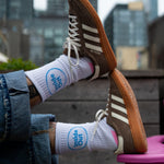 A person wearing cuffed jeans rests their feet on a pink chair, showcasing brown Adidas sneakers and white socks that feature a blue circle with the words "Inside Out."
