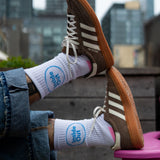 A person wearing cuffed jeans rests their feet on a pink chair, showcasing brown Adidas sneakers and white socks that feature a blue circle with the words "Inside Out."
