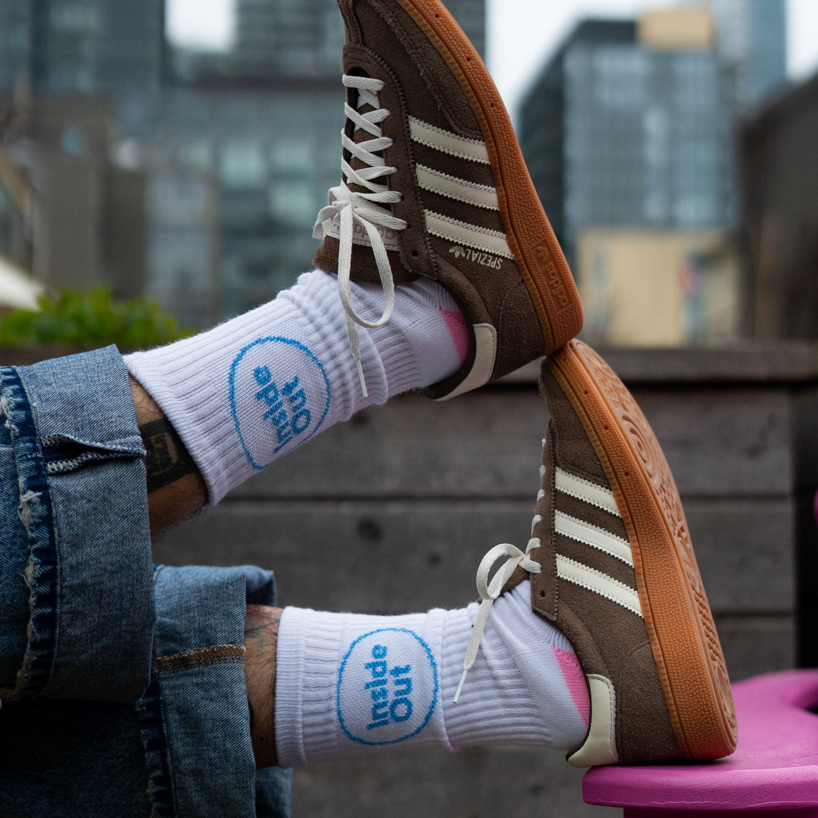 A person wearing cuffed jeans rests their feet on a pink chair, showcasing brown Adidas sneakers and white socks that feature a blue circle with the words "Inside Out."

