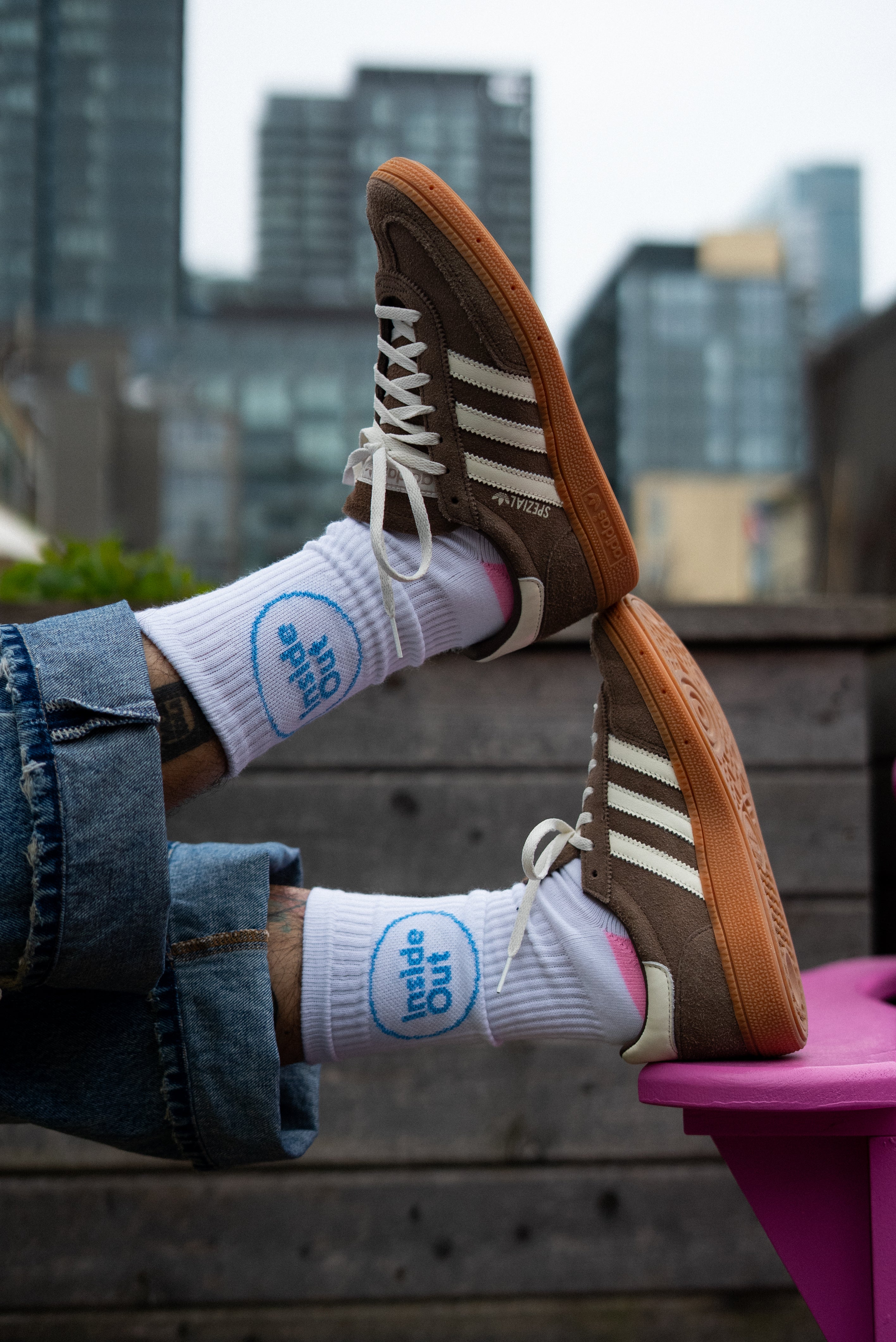 A person wearing cuffed jeans rests their feet on a pink chair, showcasing brown Adidas sneakers and white socks that feature a blue circle with the words "Inside Out."
