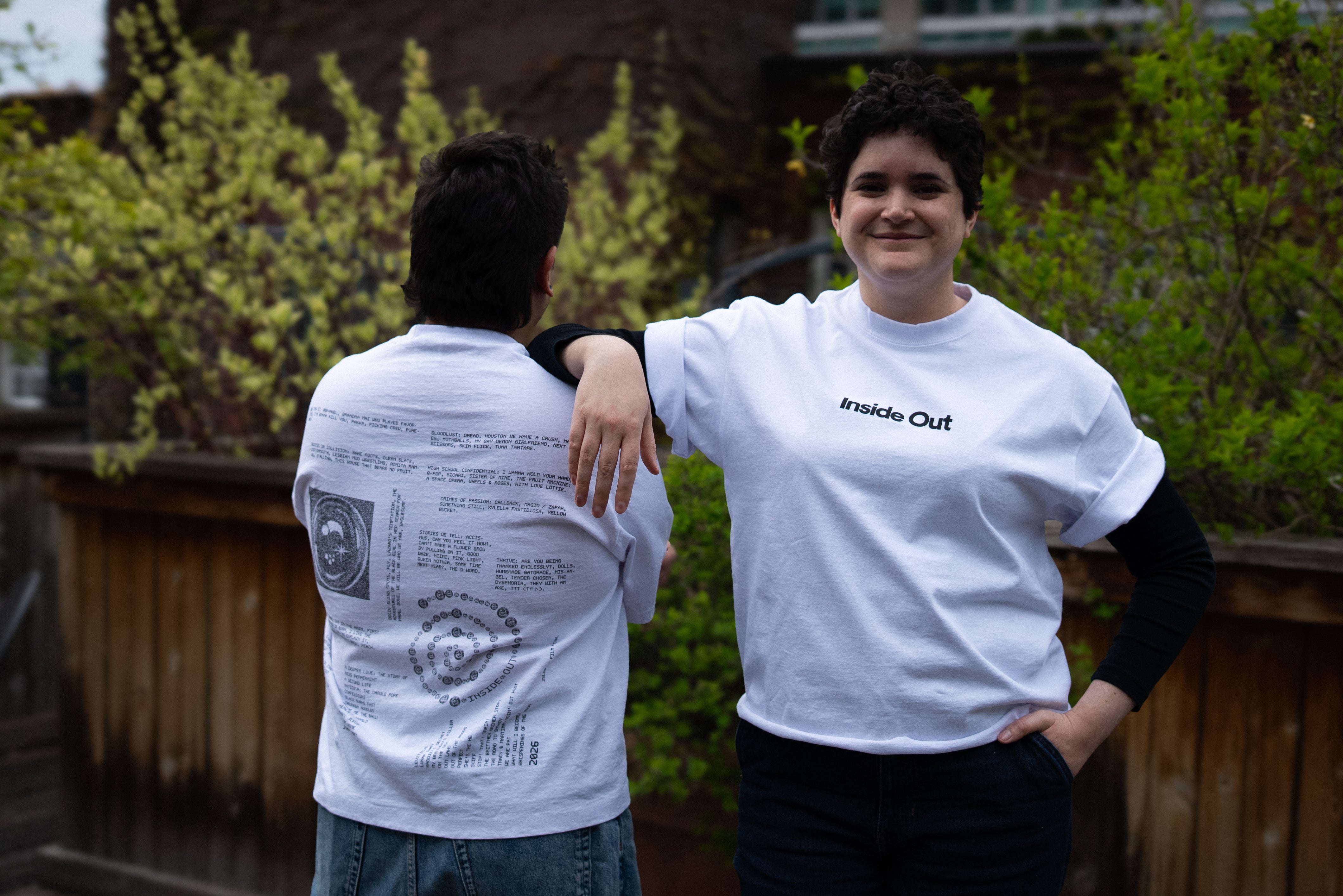 Two people lean against each other on an outdoor patio wearing matching white graphic t-shirts from "Inside Out," with one person showing the minimalist front text logo and the other showcasing a detailed back design filled with text and diagrams.