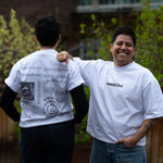 Two people lean against each other on an outdoor patio wearing matching white graphic t-shirts from "Inside Out," with one person showing the minimalist front text logo and the other showcasing a detailed back design filled with text and diagrams.