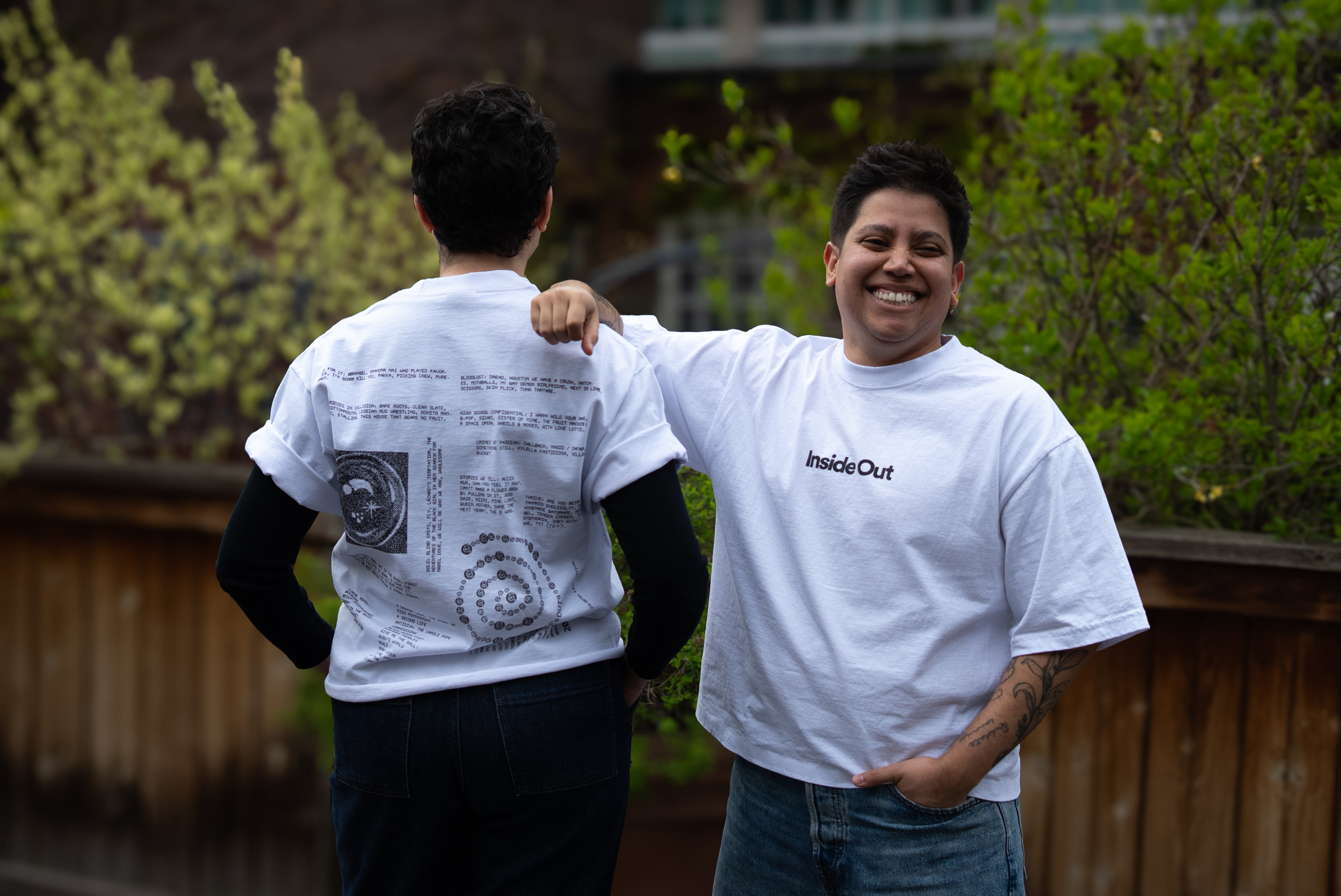 Two people lean against each other on an outdoor patio wearing matching white graphic t-shirts from "Inside Out," with one person showing the minimalist front text logo and the other showcasing a detailed back design filled with text and diagrams.
