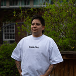 A person with arm tattoos stands outdoors with hands in their pockets, wearing a loose white t-shirt featuring the centered black text "Inside Out" and blue jeans.