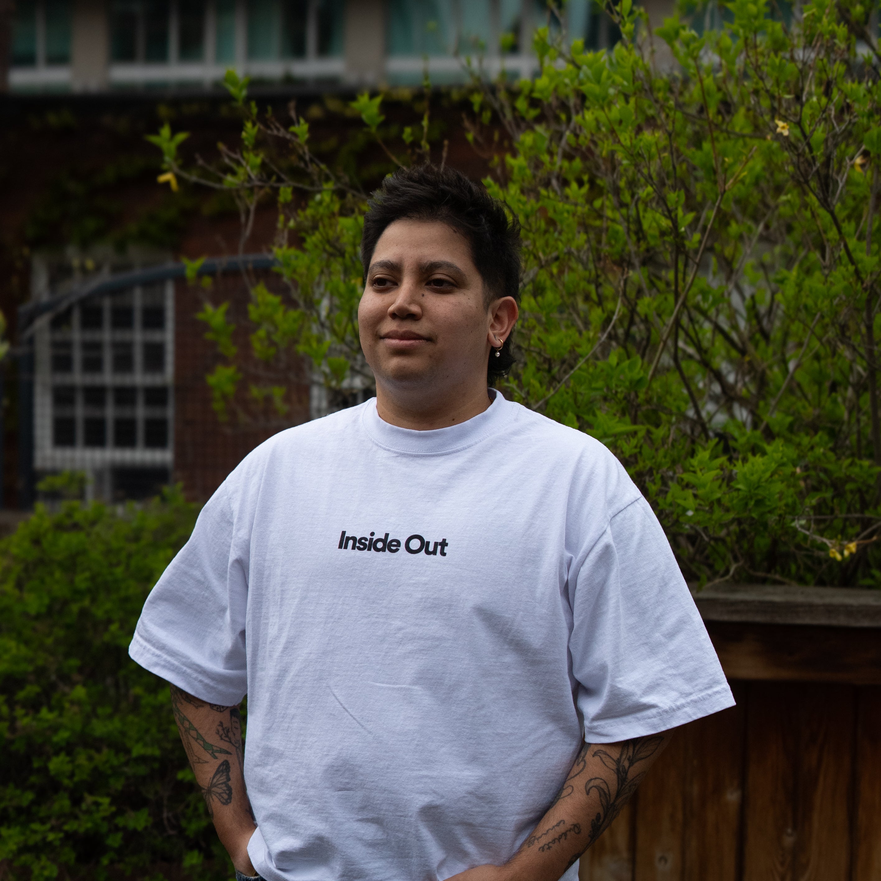 A person with arm tattoos stands outdoors with hands in their pockets, wearing a loose white t-shirt featuring the centered black text "Inside Out" and blue jeans.