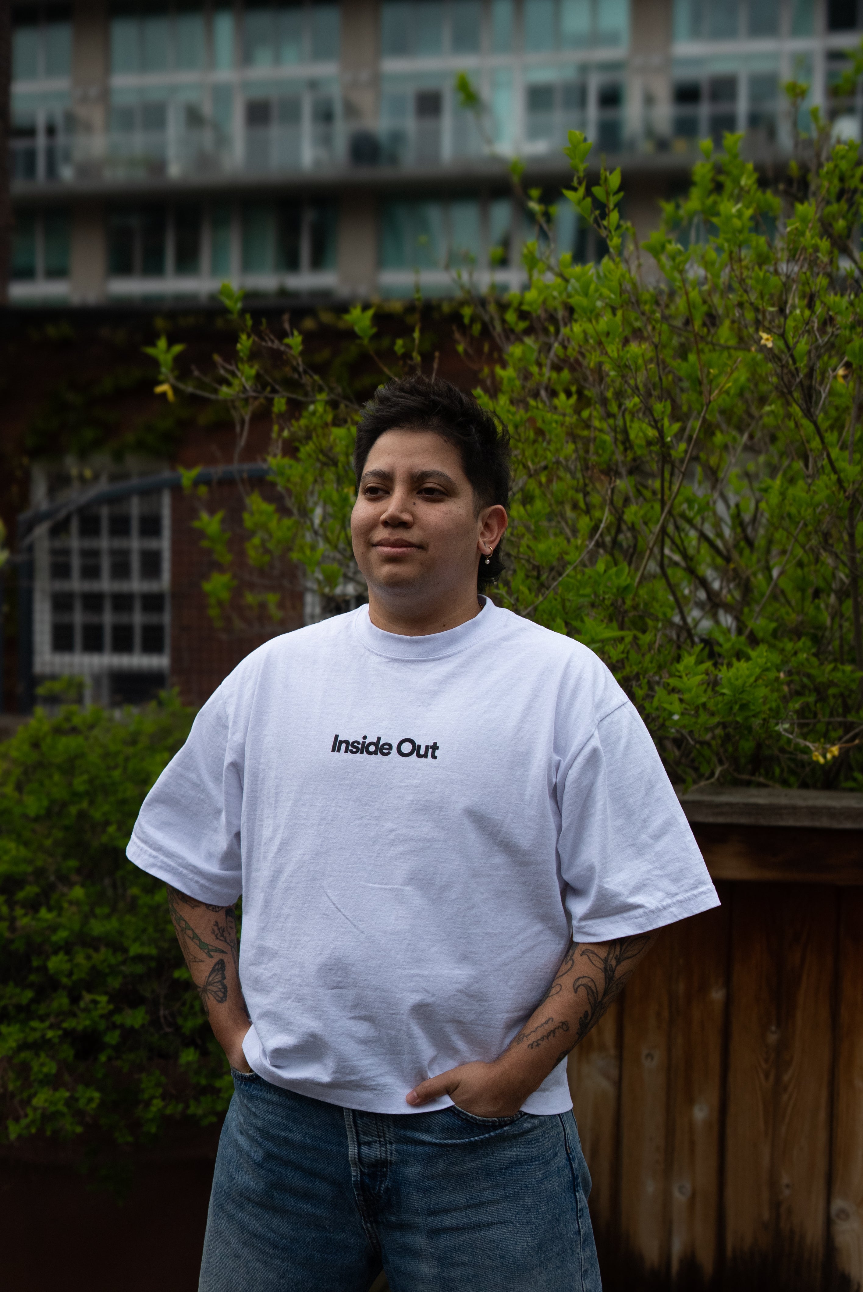 A person with arm tattoos stands outdoors with hands in their pockets, wearing a loose white t-shirt featuring the centered black text "Inside Out" and blue jeans.