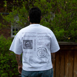 A person with short dark hair stands facing away from the camera, showcasing the back of a white t-shirt featuring intricate black text and circular diagram graphics against a backdrop of green bushes and a wooden fence.