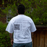 A person with short dark hair stands facing away from the camera, showcasing the back of a white t-shirt featuring intricate black text and circular diagram graphics against a backdrop of green bushes and a wooden fence.