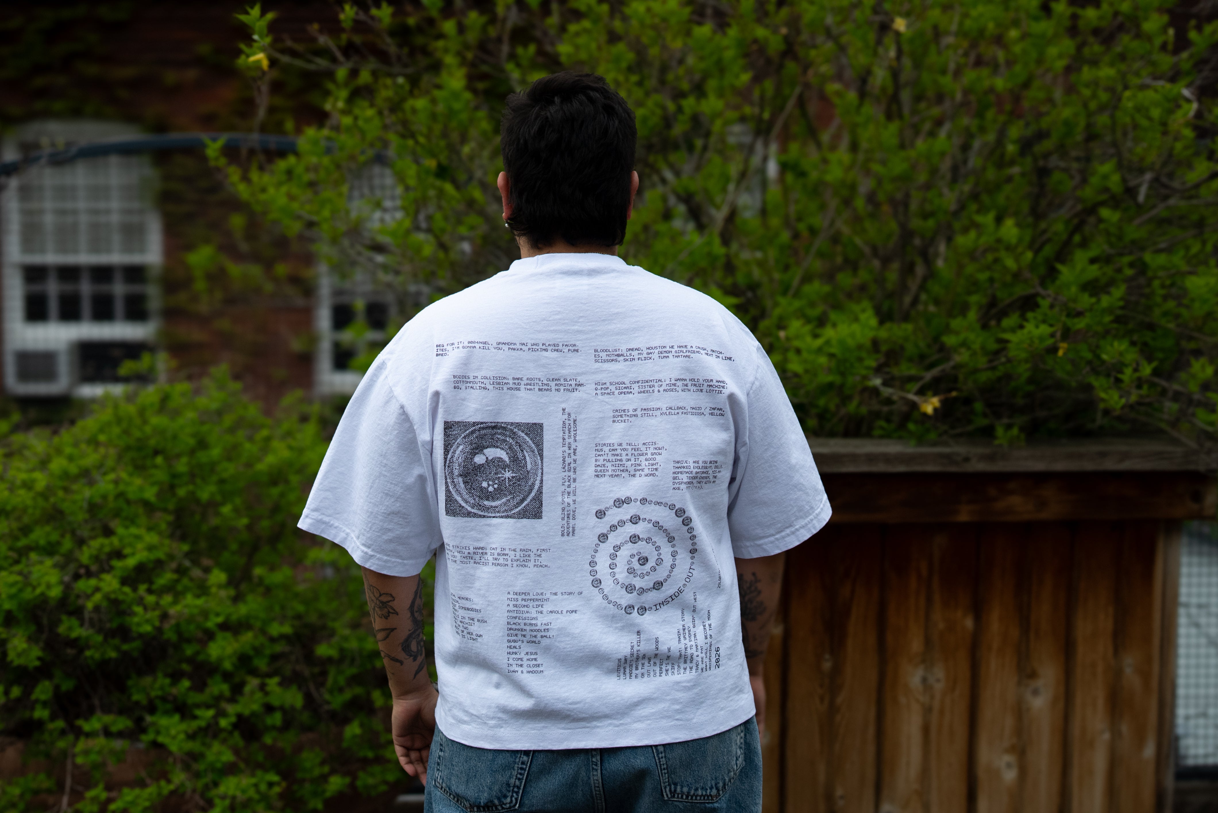 A person with short dark hair stands facing away from the camera, showcasing the back of a white t-shirt featuring intricate black text and circular diagram graphics against a backdrop of green bushes and a wooden fence.