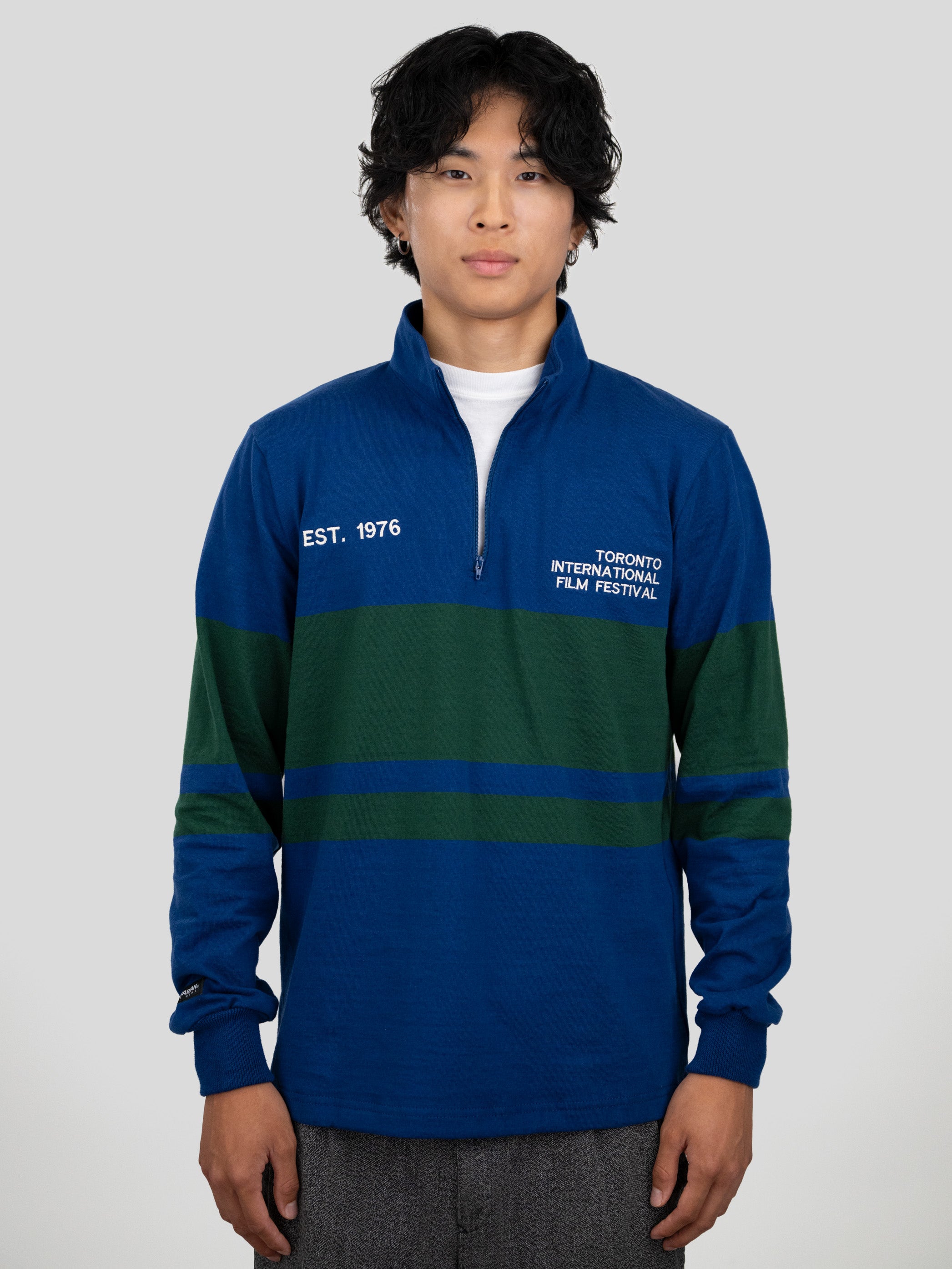 A model wears a blue and dark green quarter-zip pullover featuring "EST. 1976" and "TORONTO INTERNATIONAL FILM FESTIVAL" printed on the chest.