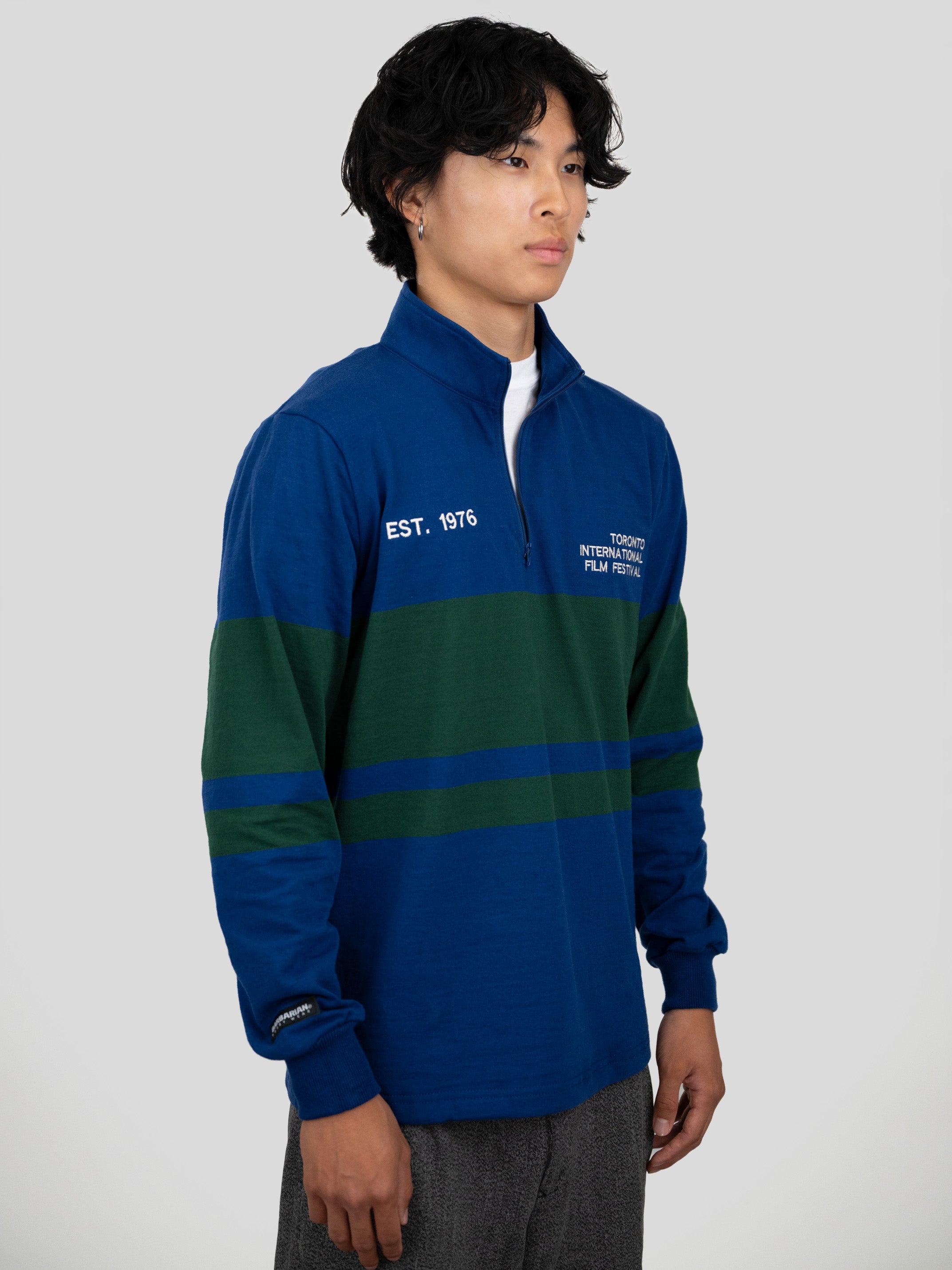 A model wears a blue and dark green quarter-zip pullover featuring "EST. 1976" and "TORONTO INTERNATIONAL FILM FESTIVAL" printed on the chest.
