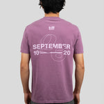 A man viewed from behind wearing a mauve-purple t-shirt with a white graphic print on the back reading 'tiff SEPTEMBER 10 20' with a ribbon design, paired with dark gray pants, against a solid light gray background.