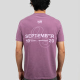 A man viewed from behind wearing a mauve-purple t-shirt with a white graphic print on the back reading 'tiff SEPTEMBER 10 20' with a ribbon design, paired with dark gray pants, against a solid light gray background.