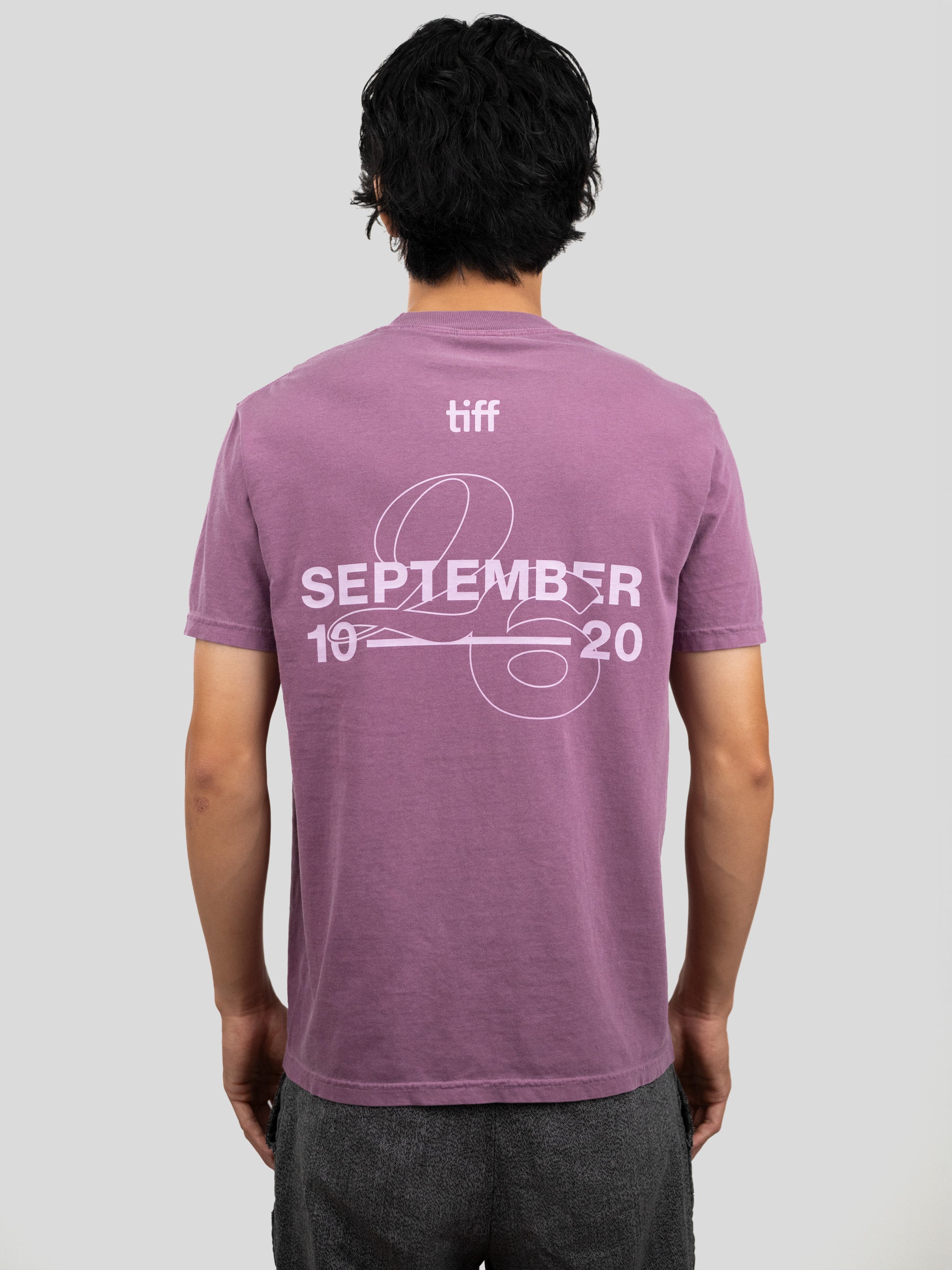 A man viewed from behind wearing a mauve-purple t-shirt with a white graphic print on the back reading 'tiff SEPTEMBER 10 20' with a ribbon design, paired with dark gray pants, against a solid light gray background.