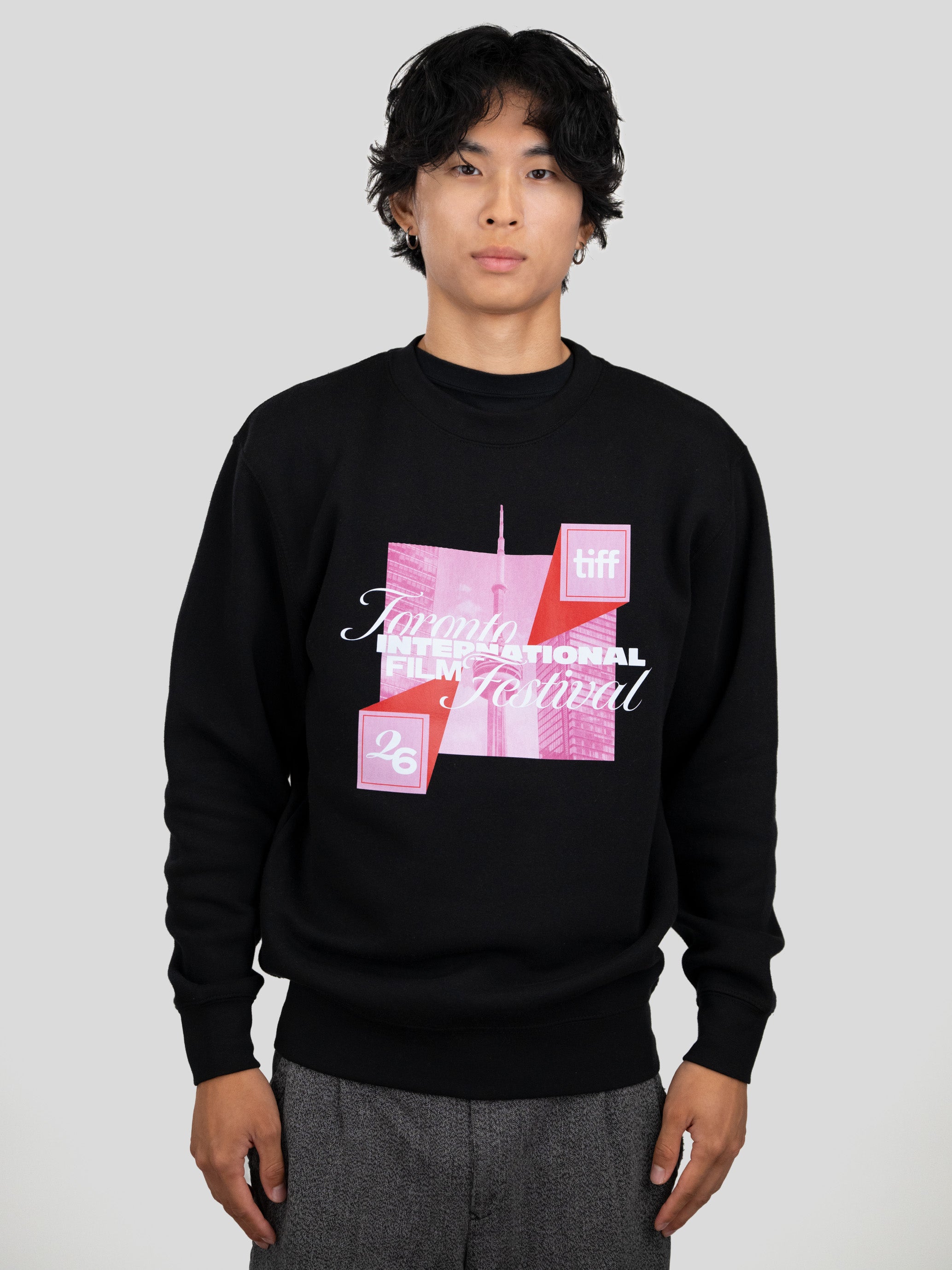 A man wearing a black crewneck sweatshirt with a pink and white graphic print for the Toronto International Film Festival, paired with gray trousers, against a solid light gray background.