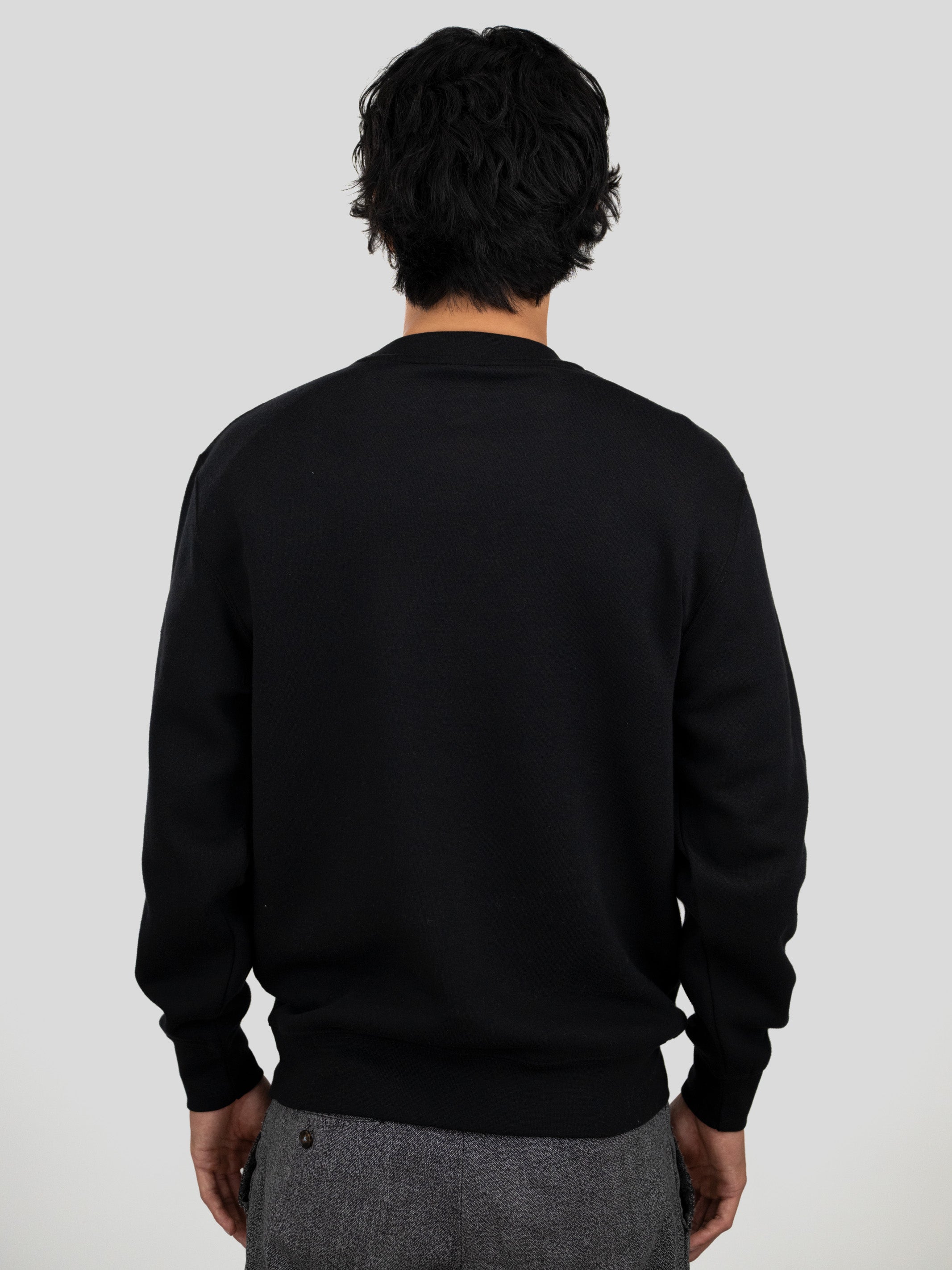 A man viewed from behind wearing a plain black long-sleeve crewneck sweater and gray trousers, set against a solid light gray background.