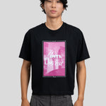 A man wearing a black short-sleeve graphic t-shirt with a pink and white 'To Intl Film Fest.' print on the front, paired with gray trousers, against a solid light gray background.