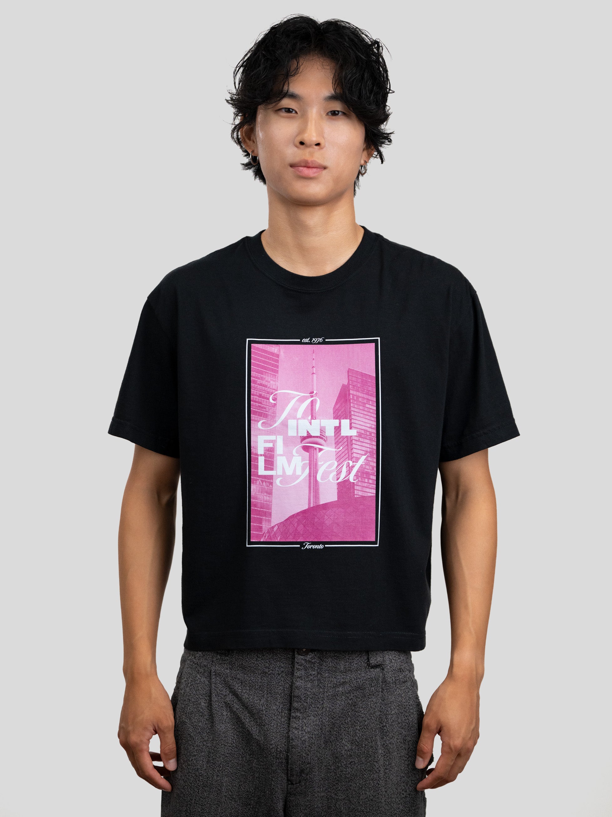 A man wearing a black short-sleeve graphic t-shirt with a pink and white 'To Intl Film Fest.' print on the front, paired with gray trousers, against a solid light gray background.
