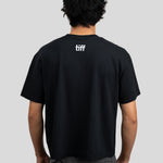 A man viewed from behind wearing a black short-sleeve T-shirt with a small white 'tiff' logo centered on the upper back, paired with gray textured pants, against a light gray studio background.