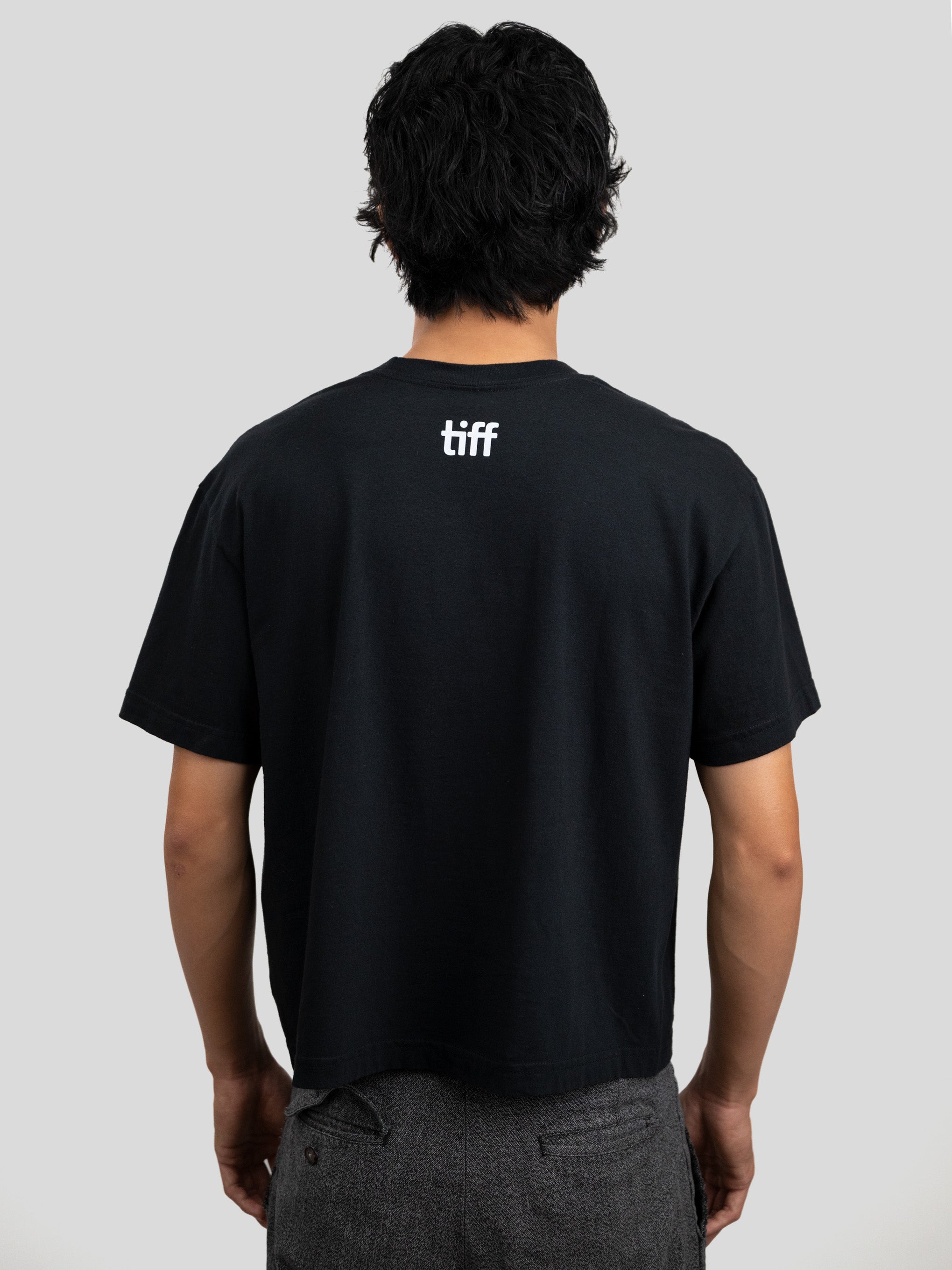 A man viewed from behind wearing a black short-sleeve T-shirt with a small white 'tiff' logo centered on the upper back, paired with gray textured pants, against a light gray studio background.