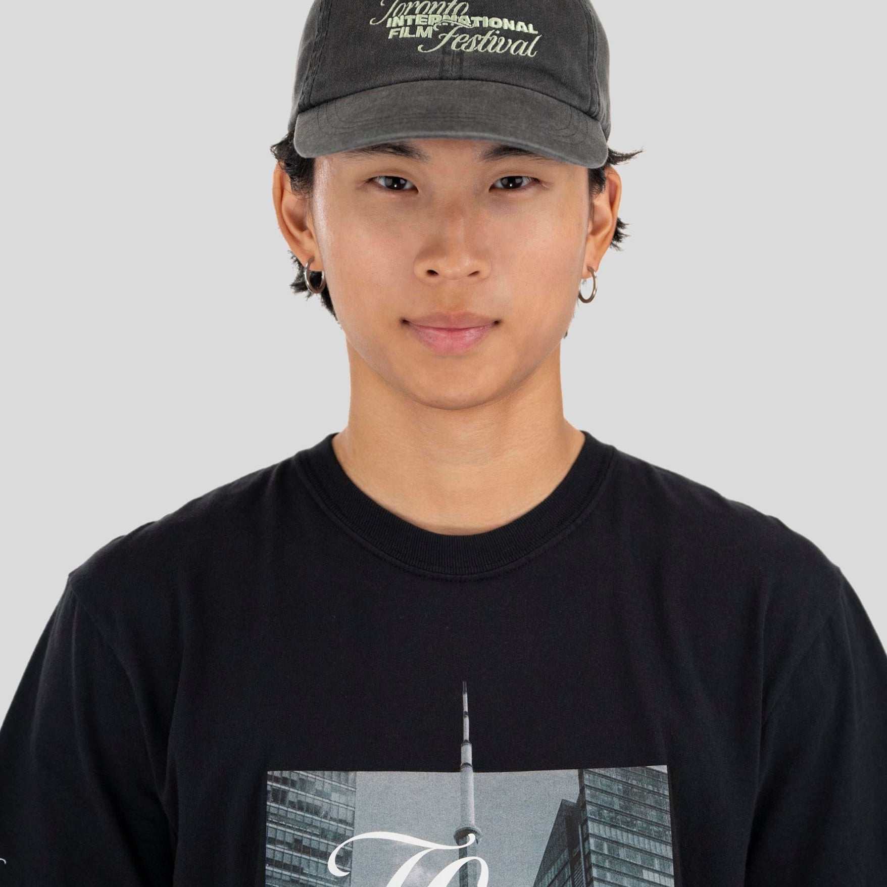 A person wearing a dark gray textured baseball cap with 'Toronto International Film Festival' embroidered on the front, paired with a black graphic t-shirt, against a solid light gray background.