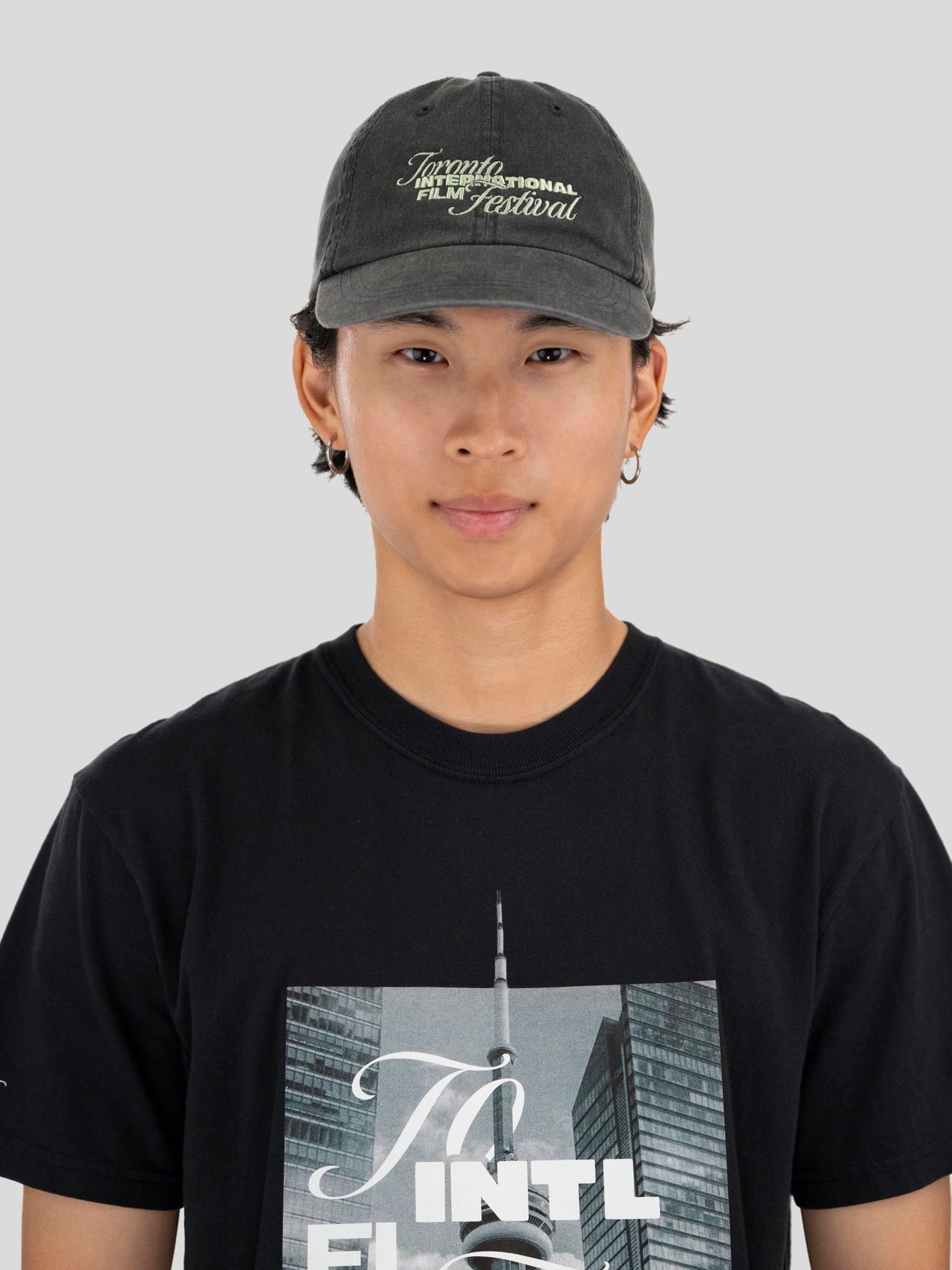 A person wearing a dark gray textured baseball cap with 'Toronto International Film Festival' embroidered on the front, paired with a black graphic t-shirt, against a solid light gray background.