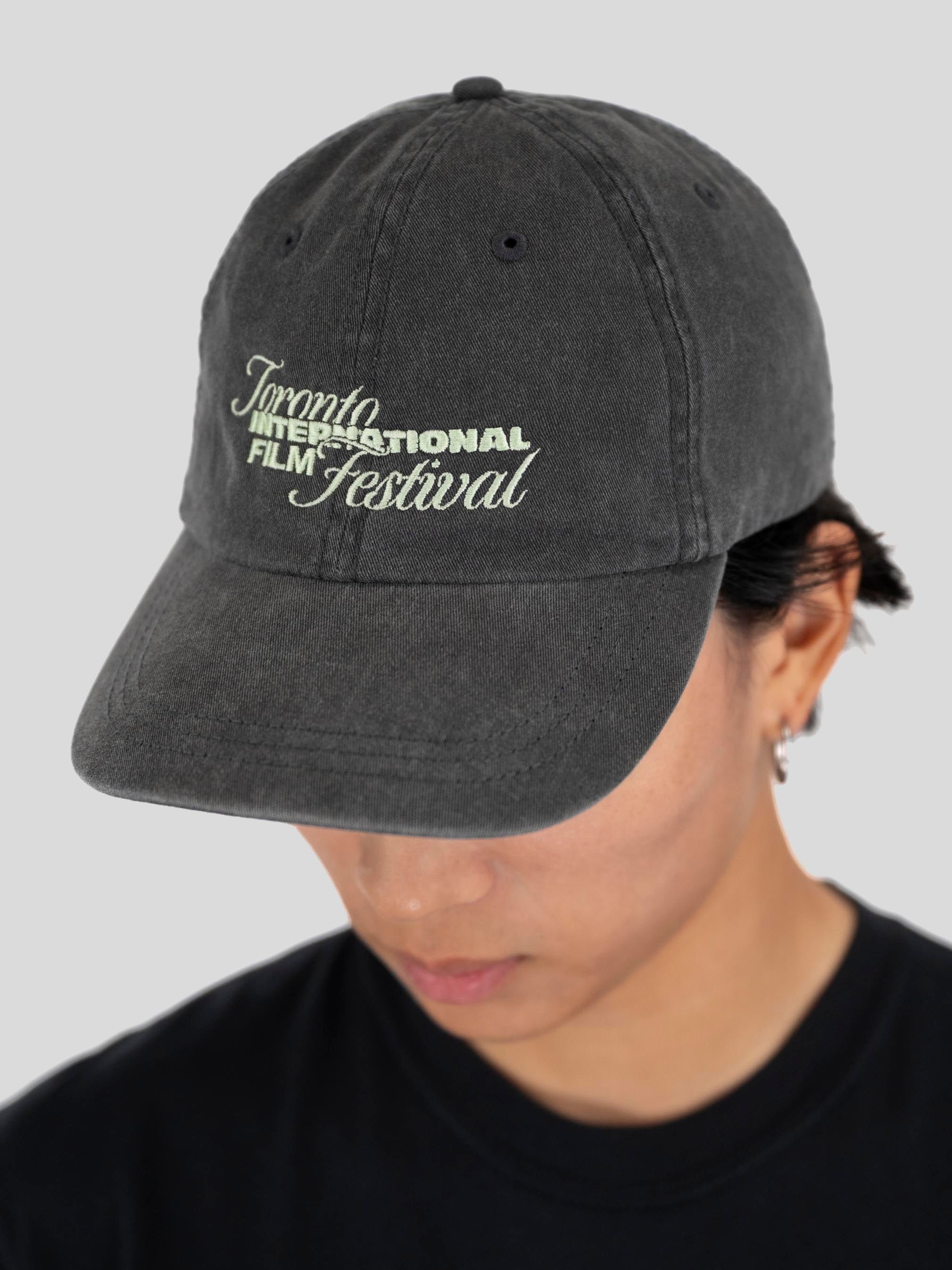 A person wearing a dark gray washed baseball cap with 'Toronto International Film Festival' embroidered in light green and white on the front, against a light gray background.