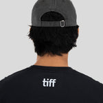 Back view of a person wearing a dark gray washed baseball cap and a black t-shirt with a white 'tiff' logo on the back, against a solid gray background.