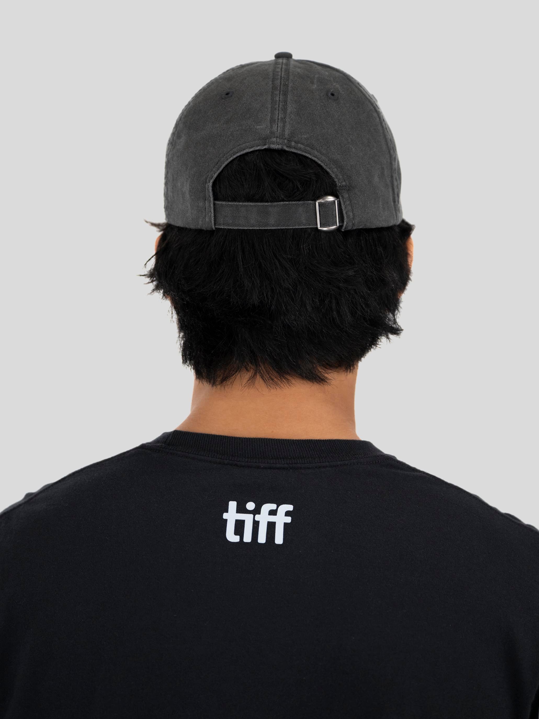 Back view of a person wearing a dark gray washed baseball cap and a black t-shirt with a white 'tiff' logo on the back, against a solid gray background.