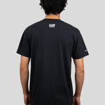 A man viewed from behind wearing a black short-sleeve T-shirt with a white 'tiff' logo printed on the upper back, paired with gray pants, against a solid light gray background.