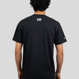 A man viewed from behind wearing a black short-sleeve T-shirt with a white 'tiff' logo printed on the upper back, paired with gray pants, against a solid light gray background.