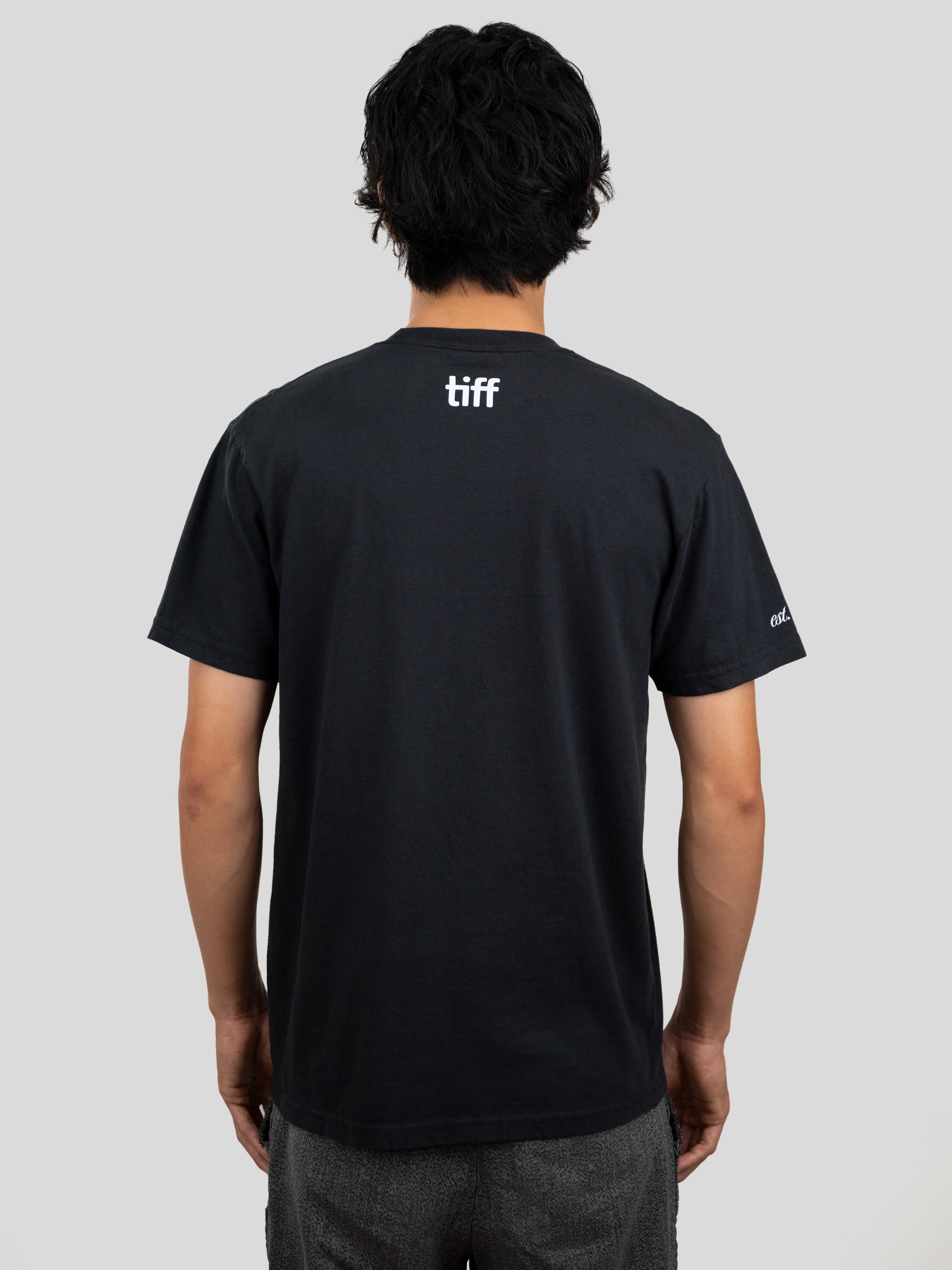 A man viewed from behind wearing a black short-sleeve T-shirt with a white 'tiff' logo printed on the upper back, paired with gray pants, against a solid light gray background.