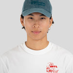 A young man wearing a dusty blue baseball cap with 'Toronto International Film Festival' embroidered on the front, paired with a white t-shirt featuring a red graphic on the chest, set against a plain light gray background.