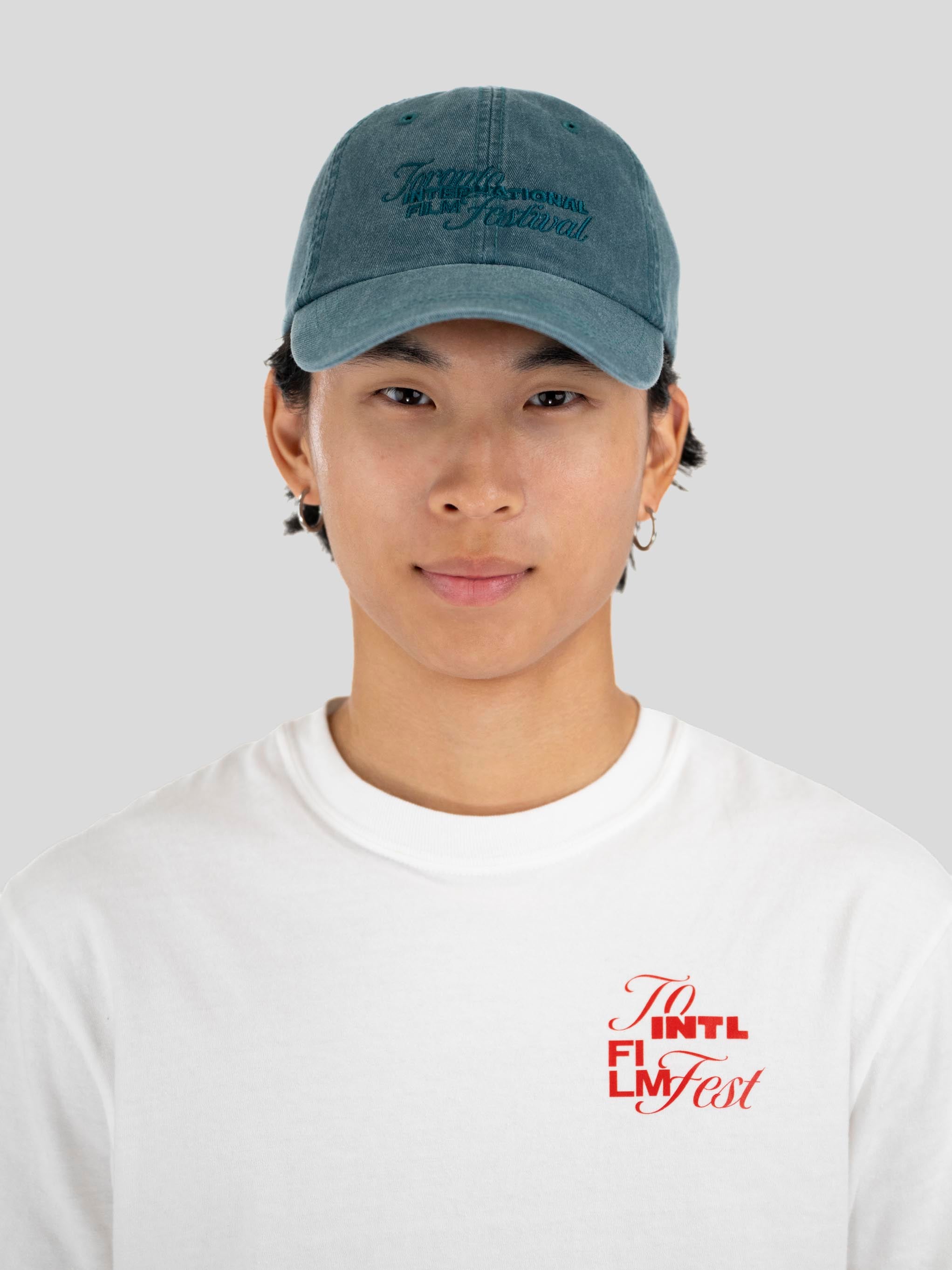 A young man wearing a dusty blue baseball cap with 'Toronto International Film Festival' embroidered on the front, paired with a white t-shirt featuring a red graphic on the chest, set against a plain light gray background.
