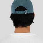 Back view of a person wearing a washed blue denim baseball cap and a white t-shirt with a red graphic print, set against a solid light gray background.