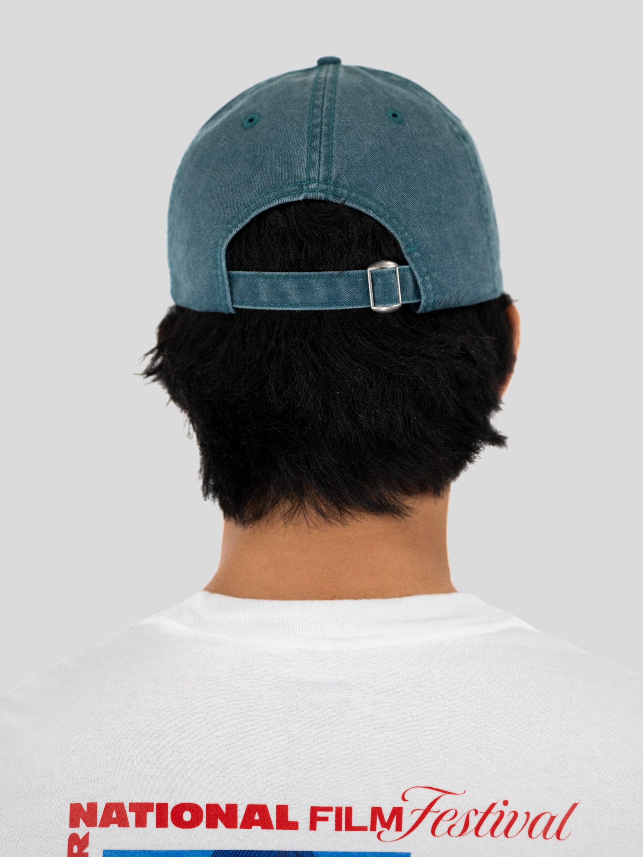 Back view of a person wearing a washed blue denim baseball cap and a white t-shirt with a red graphic print, set against a solid light gray background.