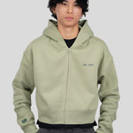 A model wearing a sage green zip-up hoodie with 'est. 1976' printed on the chest and a 'tiff' logo on the sleeve, paired with dark gray trousers, against a solid gray background.