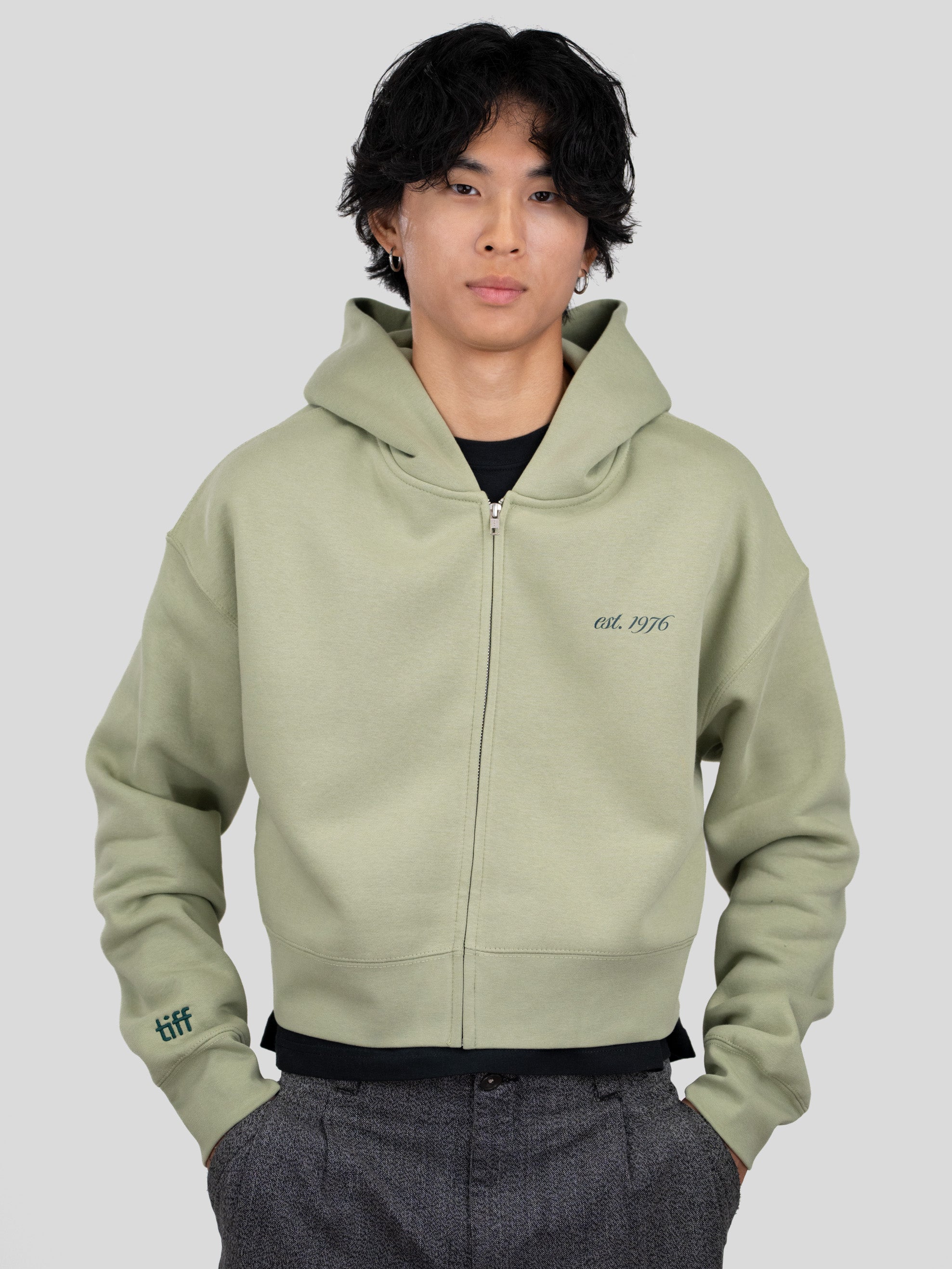 A model wearing a sage green zip-up hoodie with 'est. 1976' printed on the chest and a 'tiff' logo on the sleeve, paired with dark gray trousers, against a solid gray background.