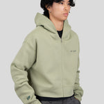 A model wearing a sage green zip-up hoodie with 'est. 1976' printed on the left chest and a 'tiff' logo on the right sleeve, paired with dark gray trousers, against a solid light gray background.